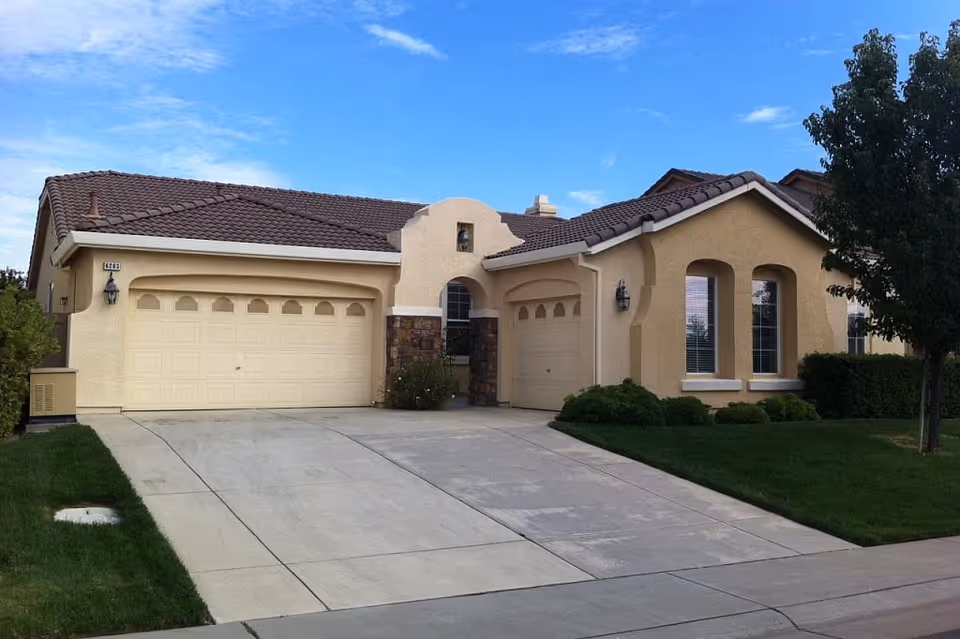 Exterior front view of a single-story residential building with beige stucco walls, a brown tiled roof, two garage doors, a driveway, and a well-maintained lawn with bushes and a tree under a blue sky with some clouds.