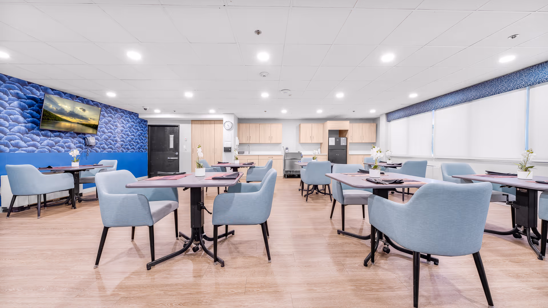 A bright and clean dining room with multiple tables and light blue upholstered chairs. Each table has a small flower arrangement and place settings. The room features a blue textured accent wall with a mounted flat-screen TV, light wood cabinetry, and large windows with white blinds and blue valances. The floor is light wood, and the ceiling has recessed lighting.