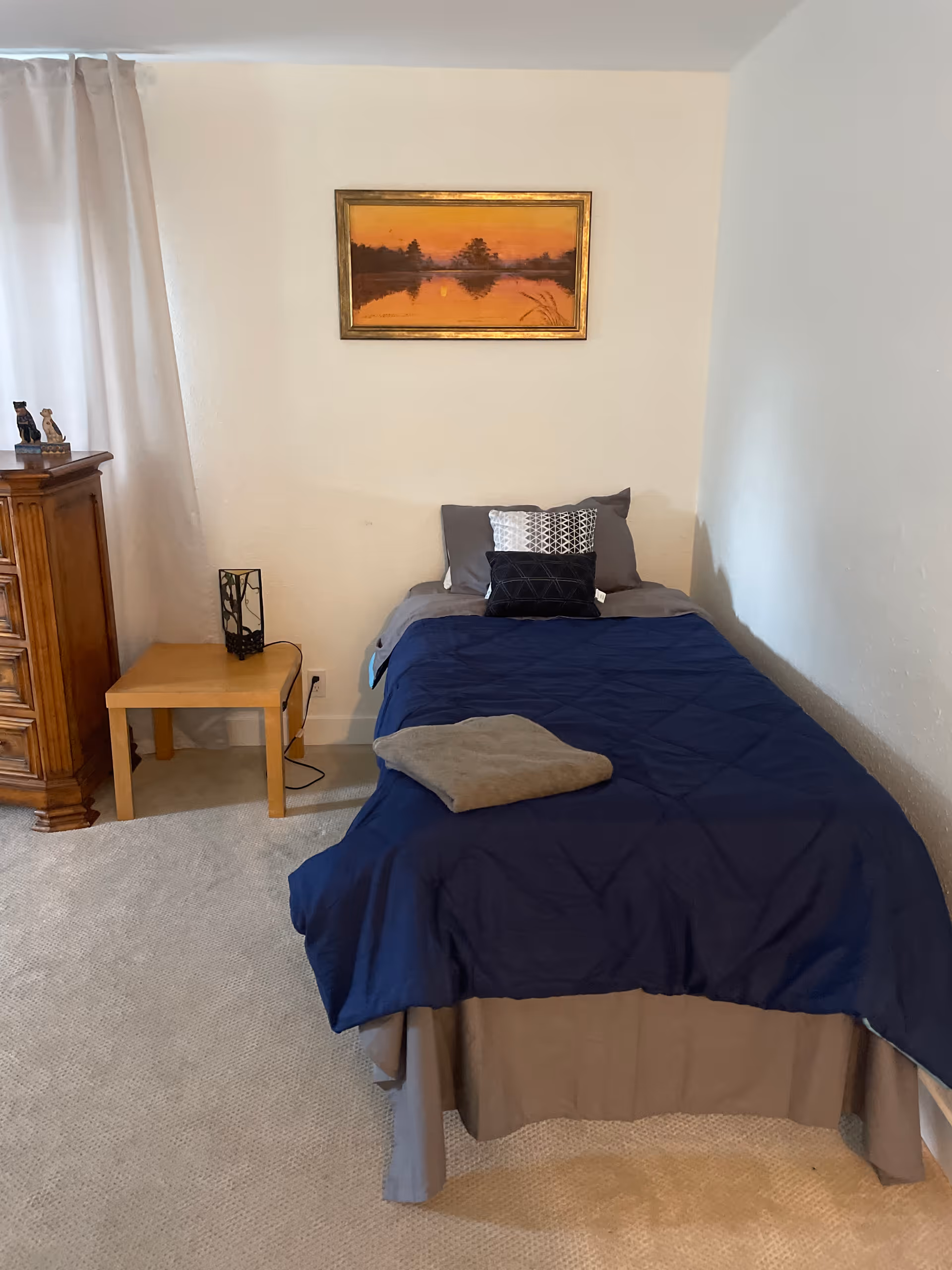 A simple bedroom with a single bed covered in a blue comforter and gray sheets, with three pillows at the head of the bed. A folded towel is placed on the bed. To the left of the bed is a wooden nightstand with a decorative lamp and a wooden dresser. A framed painting of a sunset over a lake hangs on the wall above the bed. Light-colored carpet covers the floor and a curtain is partially visible on the left side.