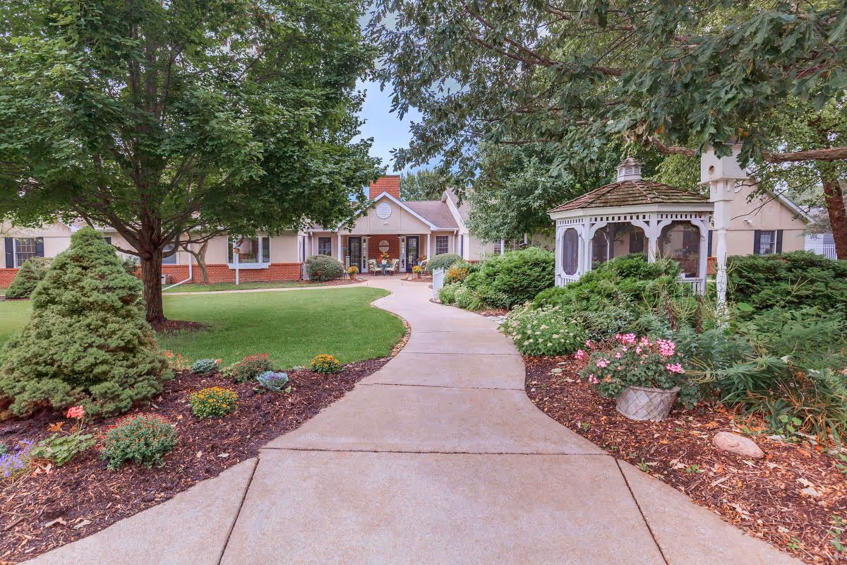 A paved walkway leads through a well-maintained garden with various shrubs, flowers, and trees towards a single-story building with a red brick base and beige walls. To the right of the path, there is a white gazebo surrounded by greenery. The sky is partly cloudy.