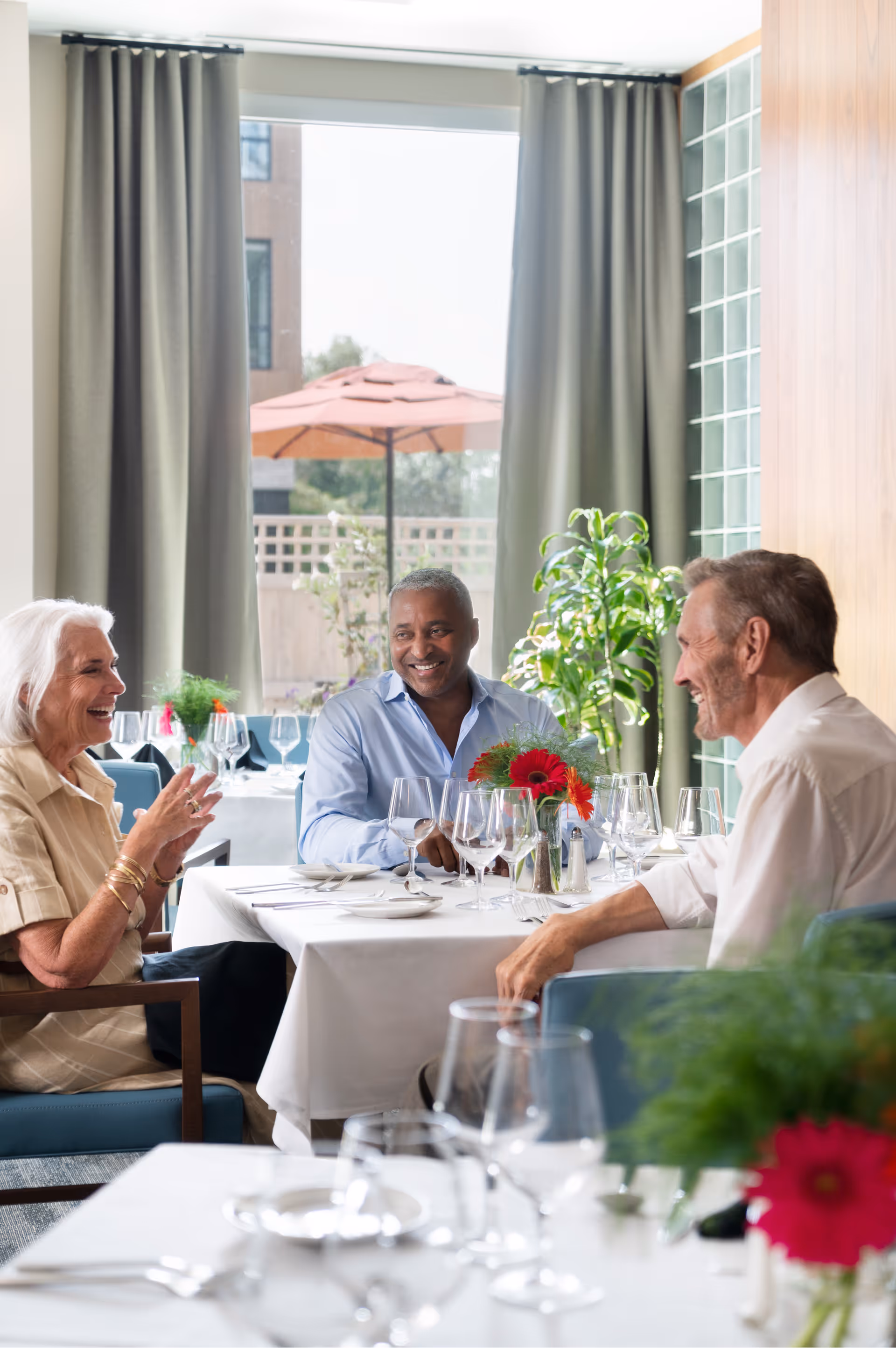 Three older adults laughing and talking at a table set with glassware and a floral centerpiece in a bright dining room.
