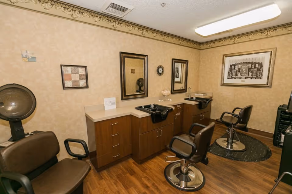 Interior view of a senior living facility's hair salon with two salon chairs in front of sinks and mirrors, a hair dryer chair, wooden cabinets, framed artwork on beige walls, and wood flooring.