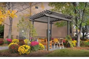 Outdoor seating area with a pergola structure surrounded by colorful flowers and bushes, located next to a brick building with trees in the background.