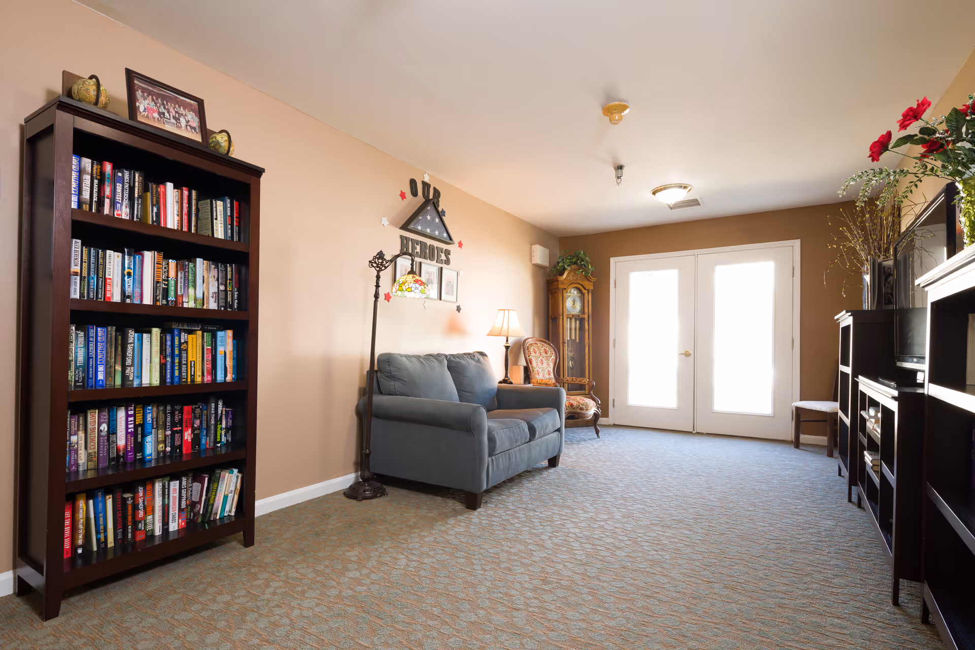 A cozy living room area with a blue loveseat, a wooden bookshelf filled with books, a vintage armchair, a grandfather clock, and a TV on a dark wooden stand. The room has beige walls, carpeted floor, and double glass doors letting in natural light. Decorative items include a floor lamp, framed pictures, and a wall decoration that reads 'Our Heroes'.