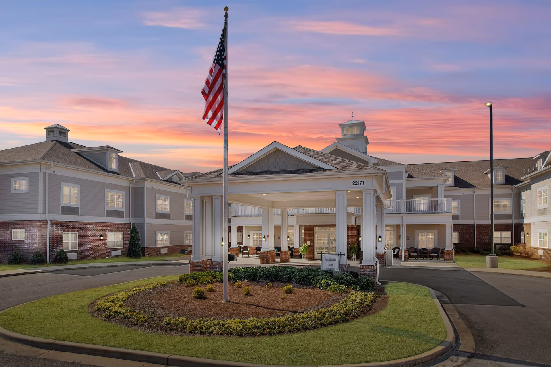 Exterior view of The Goldton at Athens senior living facility at sunset, showing a large building with a covered entrance, an American flag on a flagpole, landscaped greenery, and a colorful sky with pink and purple clouds.