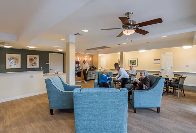 A common area in a senior living facility with several elderly people sitting and interacting. There are blue upholstered chairs arranged in a conversational grouping, a ceiling fan above, and a kitchenette area in the background where a man is standing. The room has light-colored walls, wood flooring, and framed artwork on the walls.