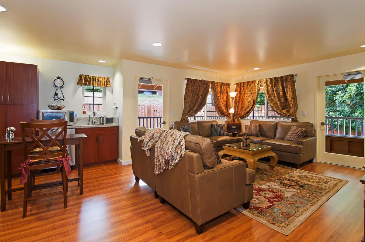 Cozy living room and kitchenette with leather sofas, a coffee table, patterned rug, wooden floors, and curtained windows.