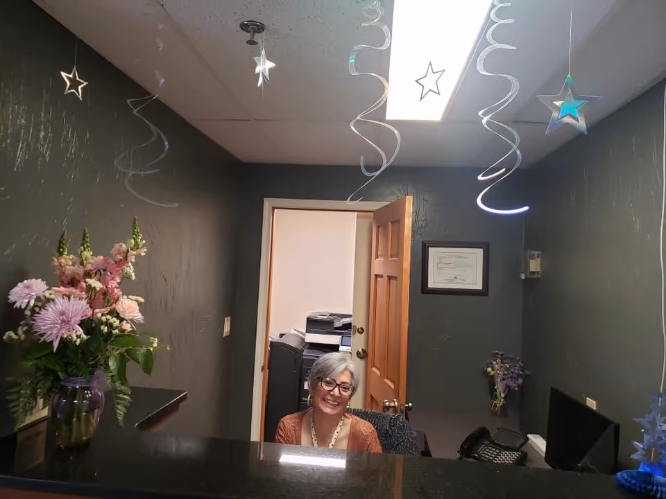 Reception area with a smiling woman sitting behind a black counter. The walls are dark gray, decorated with hanging star-shaped ornaments from the ceiling. A vase with pink and white flowers is on the left side of the counter. An open wooden door reveals an office space with a printer and other office equipment.