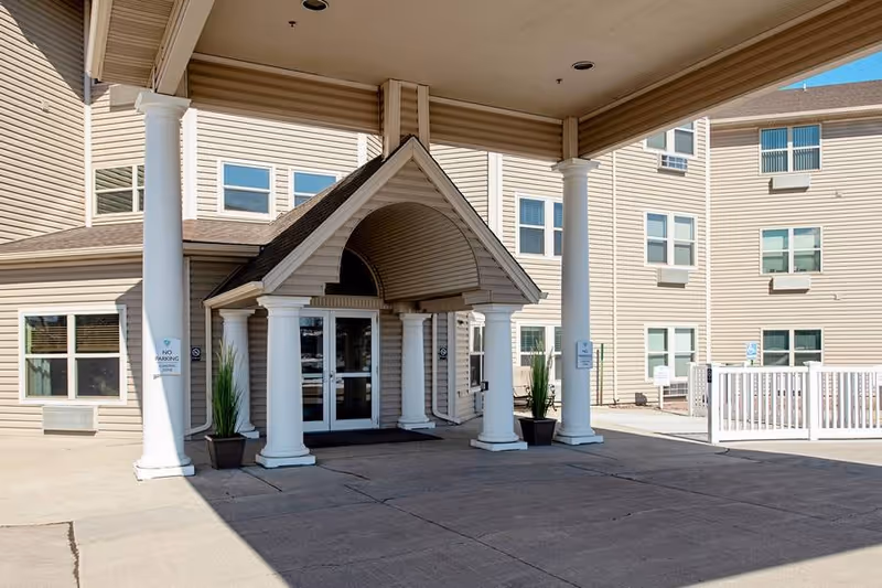 Entrance of a senior living facility with a covered driveway supported by white columns, beige siding on the building, multiple windows, and potted plants near the door.