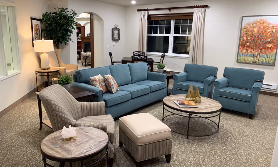 A cozy living room with a blue sofa, two matching blue armchairs, a striped armchair, and a striped ottoman. There are two round wooden coffee tables, one with decorative items and magazines. The room has beige carpet, a large window with beige curtains, a colorful painting of trees on the wall, a side table with a lamp, and a potted plant. In the background, there is a dining table with chairs and an archway leading to another room.