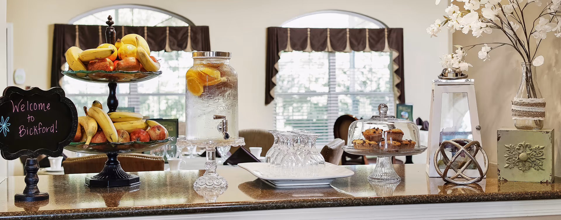 A countertop display in a dining or common area featuring a two-tiered fruit stand with bananas and apples, a glass dispenser filled with water and lemon slices, a tray with upside-down glasses, a cake stand with muffins under a glass cover, and decorative items including a vase with white flowers and a small chalkboard sign that reads 'Welcome to Bickford!'. In the background, there are windows with brown valances and a glimpse of dining tables and chairs.