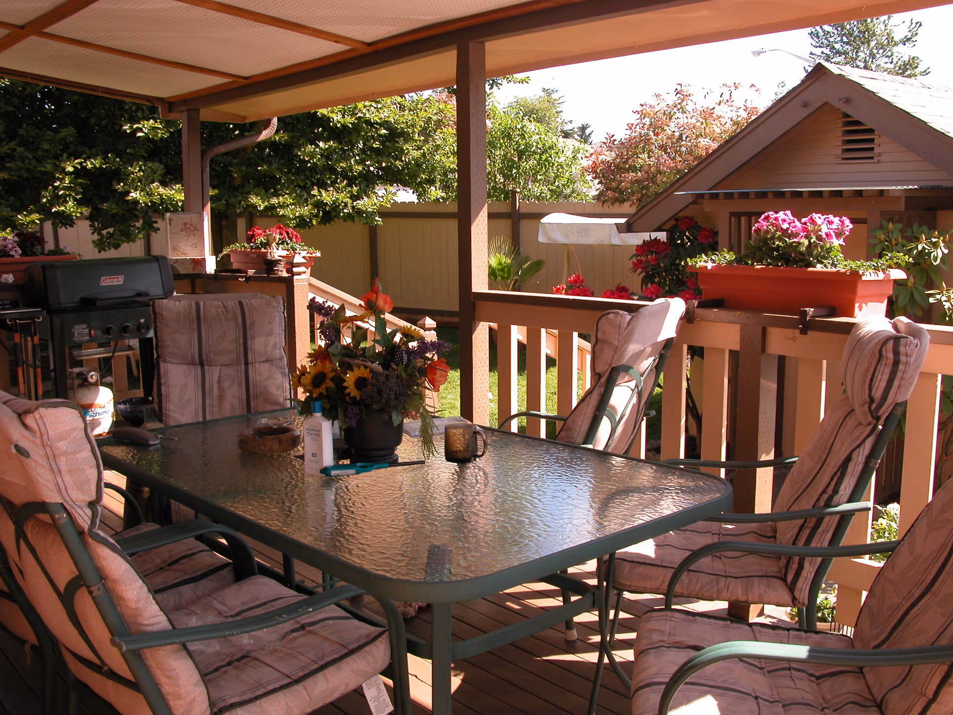 Covered outdoor patio with a glass-top table surrounded by six cushioned chairs. The table has a flower arrangement, a mug, and some small items on it. There is a barbecue grill in the corner and flower pots with blooming flowers on the railing. Trees and a small shed are visible in the background.