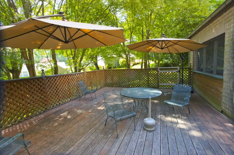 Outdoor wooden deck area with metal chairs and round tables shaded by large beige umbrellas, surrounded by lattice fencing and green trees in the background.
