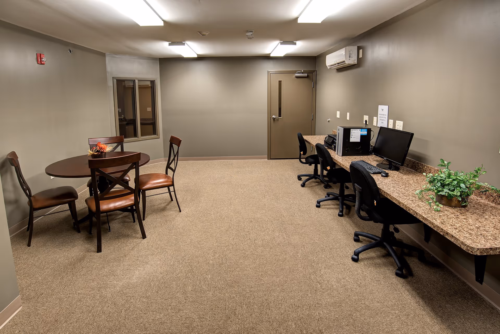 A room with beige walls and carpeted floor featuring a round wooden table with four chairs on the left side and a long counter with three office chairs, a computer, a printer, and a potted plant on the right side. There is a door and a window on the far wall, and fluorescent ceiling lights illuminate the space.