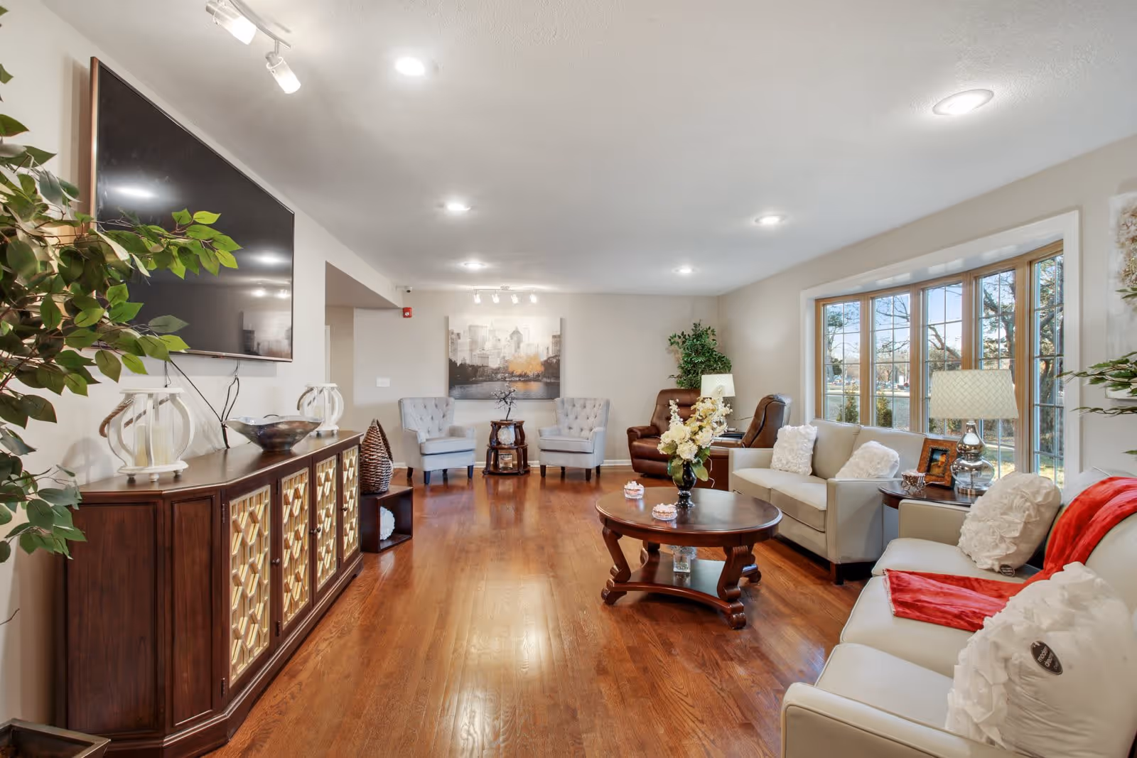 A spacious and well-lit living room with wooden flooring, featuring a large flat-screen TV mounted on the wall above a wooden cabinet with decorative panels. The room has multiple seating options including two beige sofas with white pillows and a red throw, two gray armchairs, and a brown recliner. A round wooden coffee table with a flower vase and small decorative items is centered in the room. Large windows on the right side allow natural light to fill the space, and there are several plants and lamps adding to the cozy atmosphere.