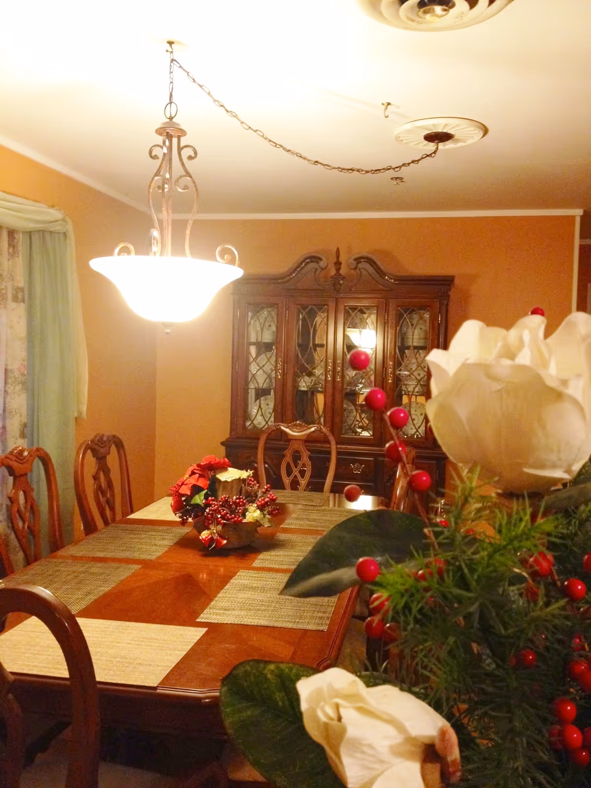 A warmly lit dining room with a wooden dining table set with placemats and a festive centerpiece featuring red berries and greenery. There are six wooden chairs around the table. In the background, there is a wooden china cabinet with glass doors displaying dishes. A hanging light fixture with a frosted glass shade illuminates the room. A window with green curtains and floral drapes is visible on the left side.