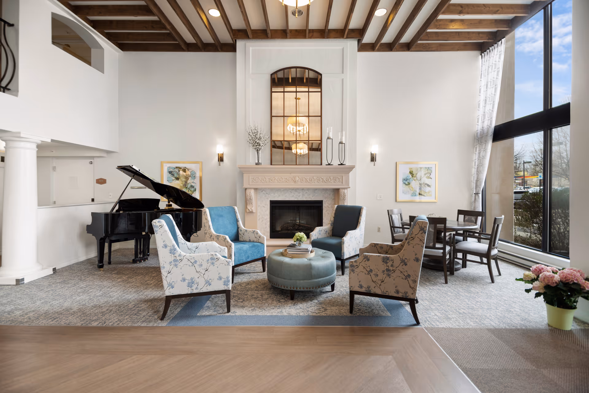 A bright and elegant living room area with a grand piano on the left, four upholstered armchairs arranged around a round ottoman in the center, a fireplace with a large mirror above it, and a dining table with chairs near large floor-to-ceiling windows letting in natural light. The ceiling features exposed wooden beams and there are decorative wall sconces and framed artwork on the walls.