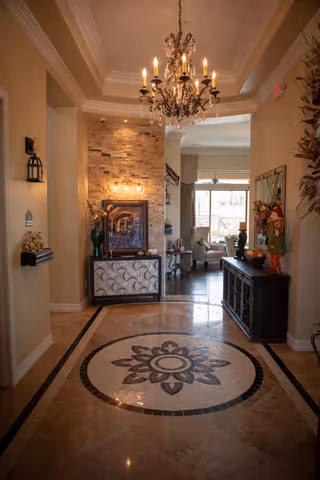A warmly lit hallway with a decorative chandelier hanging from the ceiling. The floor features a circular floral mosaic design. On the left side, there is a small shelf with decorative items and a wall-mounted lantern. At the end of the hallway, there is a stone accent wall with a framed picture and a cabinet beneath it. To the right, a dark wooden sideboard with floral arrangements and other decor items is visible. The hallway opens into a bright living area with large windows and seating.