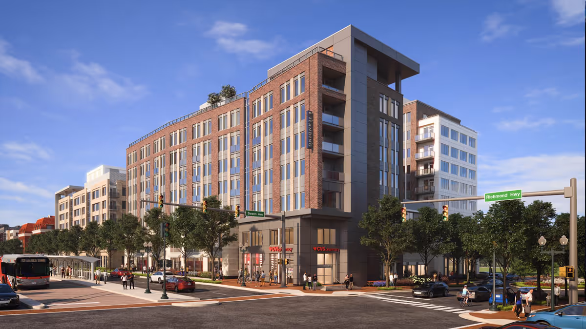 Street view of a modern multi-story brick and glass building on a sunny day, with trees lining the sidewalks and people walking near a bus stop. The building has a CVS Pharmacy on the ground floor and street signs indicating the intersection of Richmond Hwy and Swann Ave.