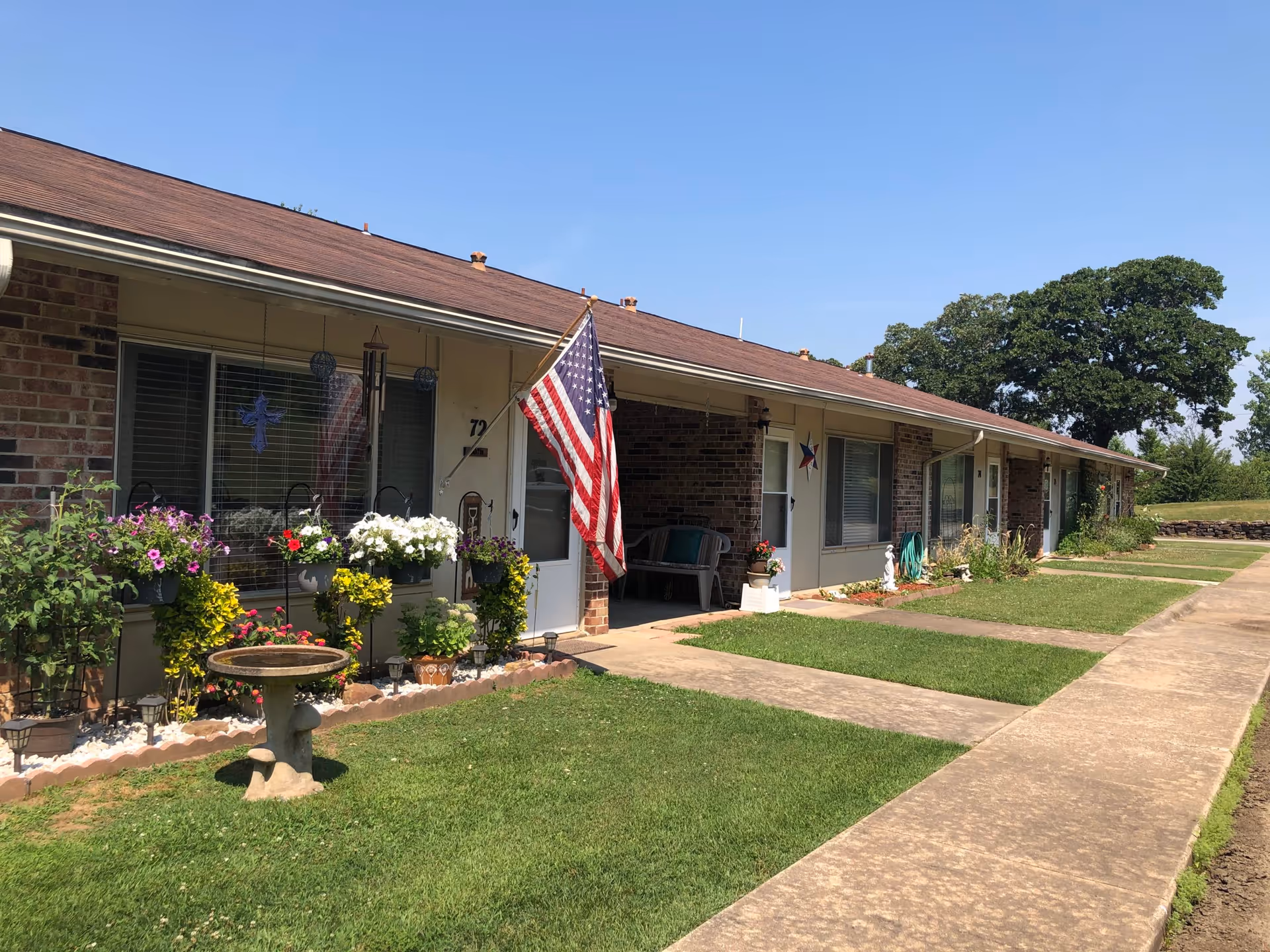 Exterior view of a single-story brick building with several doors and windows, each with small garden areas in front. An American flag is displayed near one of the doors, and there is a birdbath on the lawn. The sky is clear and blue.