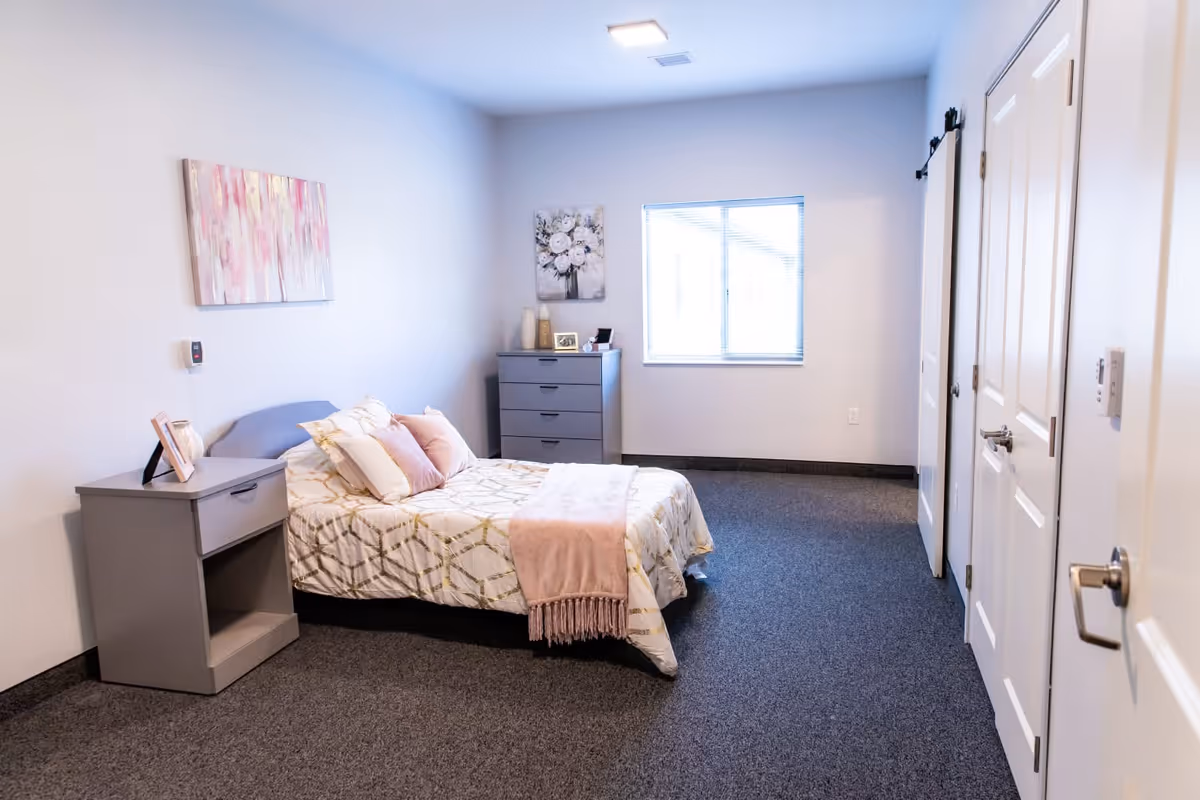 A simple, clean bedroom with a bed featuring white and gold patterned bedding and pink pillows. There is a gray nightstand with a framed photo and a gray dresser against the far wall. Two pieces of floral artwork hang on the walls, and a window lets in natural light. The room has gray carpet and white walls with a closed door on the right side.