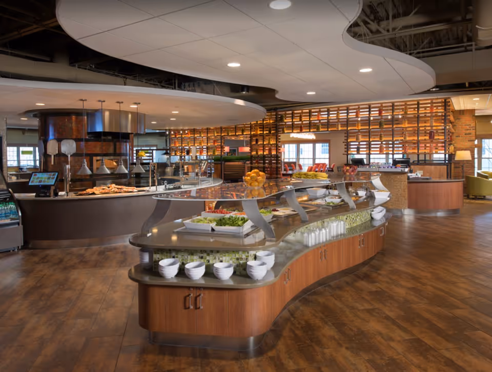 A spacious and modern dining area with a curved buffet counter displaying various fresh foods including salads and fruits. The room features warm wood flooring, contemporary lighting, and a pizza oven area in the background. There are shelves with decorative items and seating areas visible further back.