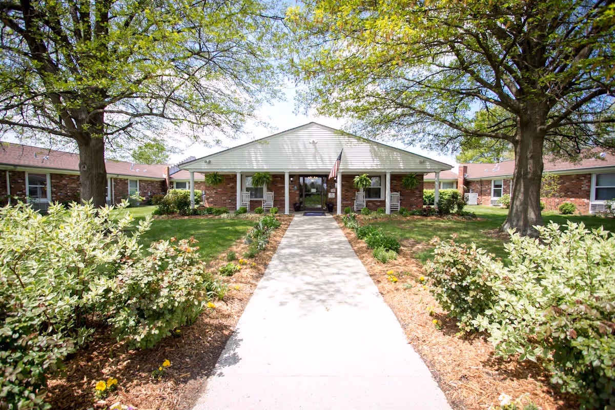 Front exterior view of Bluegrass Care & Rehabilitation building with a concrete walkway leading to the entrance. The building is single-story with brick walls and a white gabled roof. There are two large trees and landscaped bushes and flowers on either side of the walkway. The entrance has a covered porch with rocking chairs and hanging plants, and an American flag is displayed near the door.