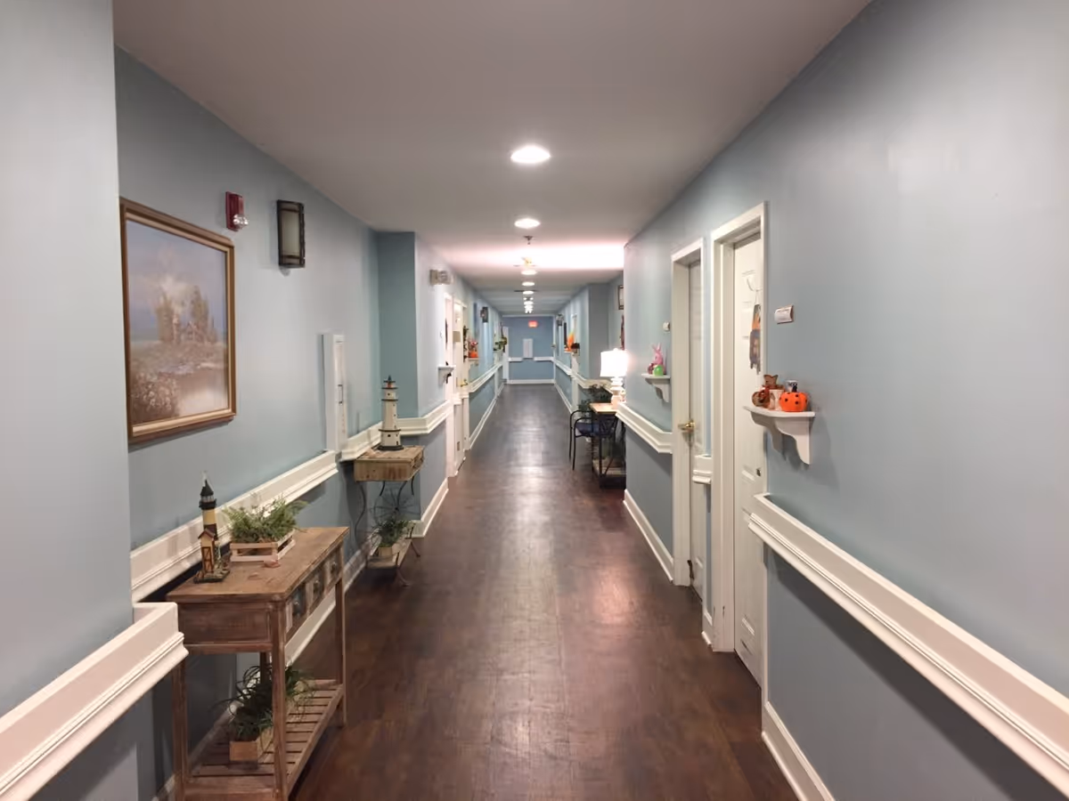 A long, well-lit hallway in an assisted living facility with light blue walls, dark wood flooring, and white trim. The hallway features small tables and shelves decorated with plants, lighthouse figurines, and seasonal decorations. Several closed white doors line the hallway on both sides.