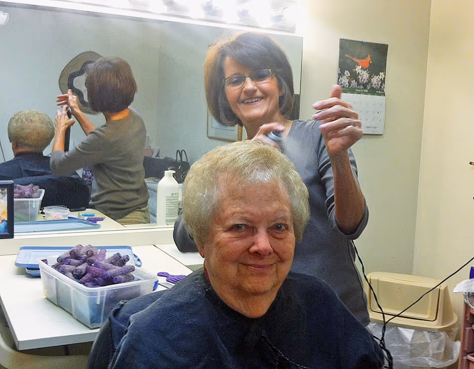 An elderly woman with short blonde hair sits in a salon chair while a hairstylist sprays her hair. The hairstylist is smiling and standing behind her. The room has a large mirror reflecting both women, a calendar on the wall, and a container of purple hair rollers on the counter.