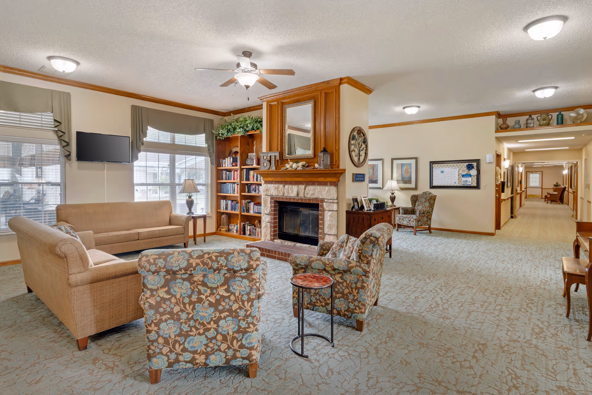 A cozy living room area in a senior living facility featuring a stone fireplace with a wooden mantel and mirror above it. The room has two floral-patterned armchairs, a beige sofa, a small round side table, a wooden bookshelf filled with books, and large windows with valances allowing natural light to enter. The walls are decorated with framed artwork and a bulletin board, and the carpet has a subtle pattern. Ceiling lights and a ceiling fan provide illumination.