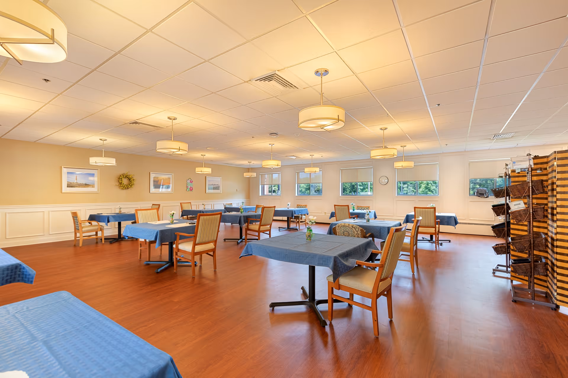 Spacious dining room with multiple tables covered in blue tablecloths, wooden chairs, pendant lights, and windows along the far wall.