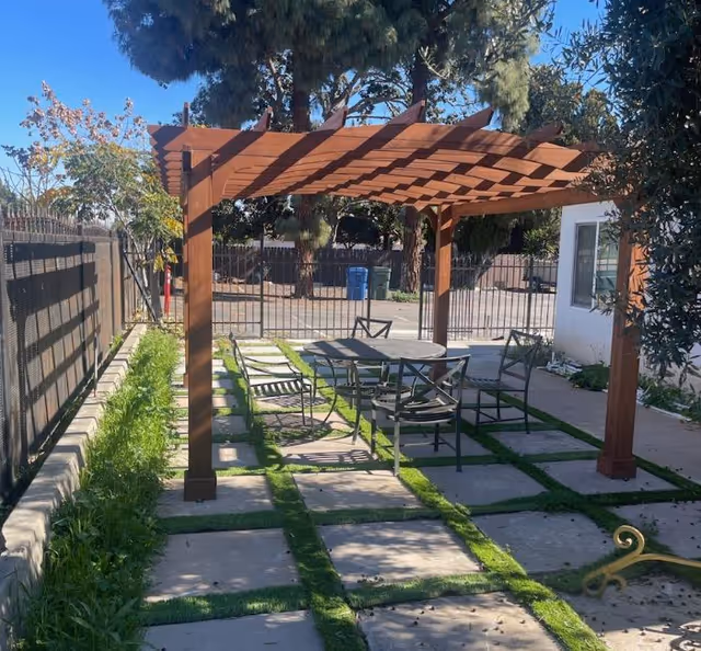 Wooden pergola shading a patio with a table and chairs on a grid of pavers and grass in an outdoor courtyard.