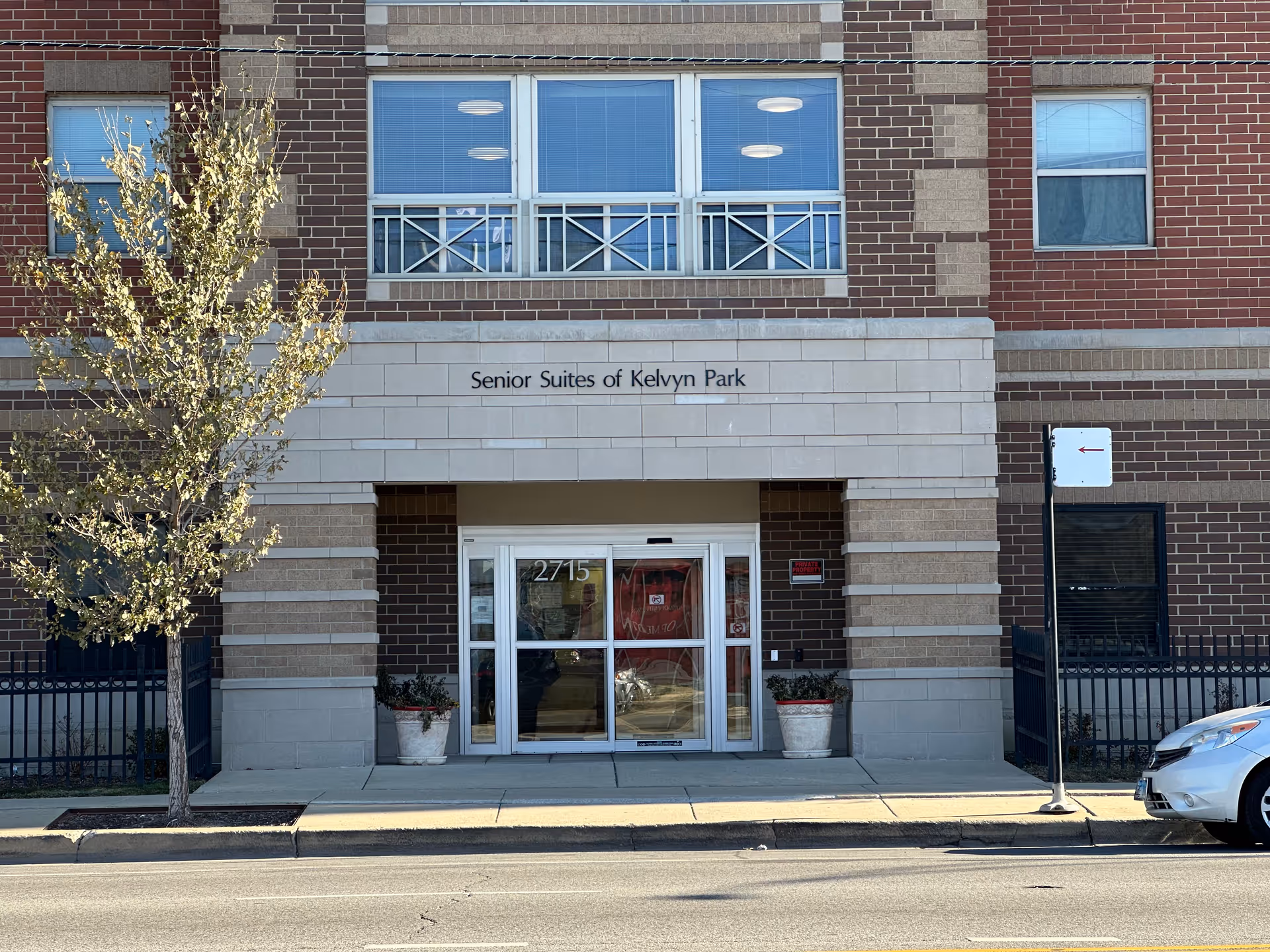 Front entrance of a brick building labeled 'Senior Suites of Kelvyn Park' with glass doors and planters at the sidewalk.