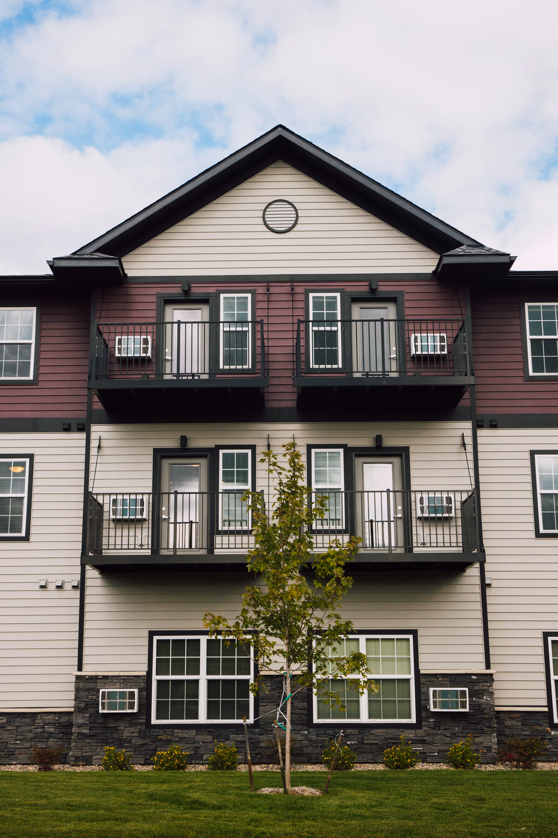 Exterior view of a multi-story residential building with balconies, multiple windows, and a small tree in front on a grassy lawn under a partly cloudy sky.