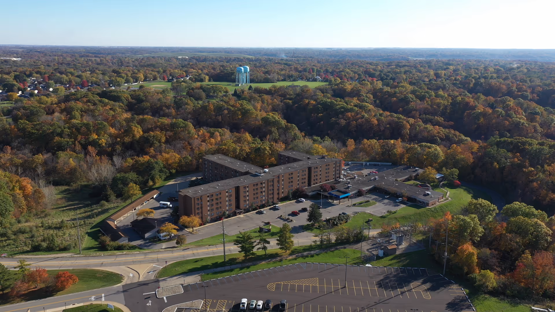 Aerial view of Saxony Court Senior Living facility surrounded by dense trees with autumn foliage. The multi-story building has a large parking lot with several cars parked. In the background, there are water towers and a vast expanse of forested land under a clear sky.