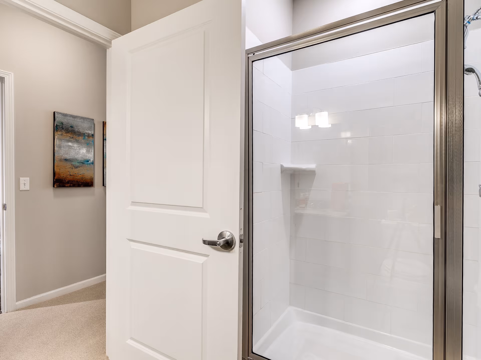 View of a bathroom shower with a glass door and white tiled walls, seen through an open white door. A hallway with beige walls and carpet is visible outside the bathroom, along with a piece of abstract wall art.