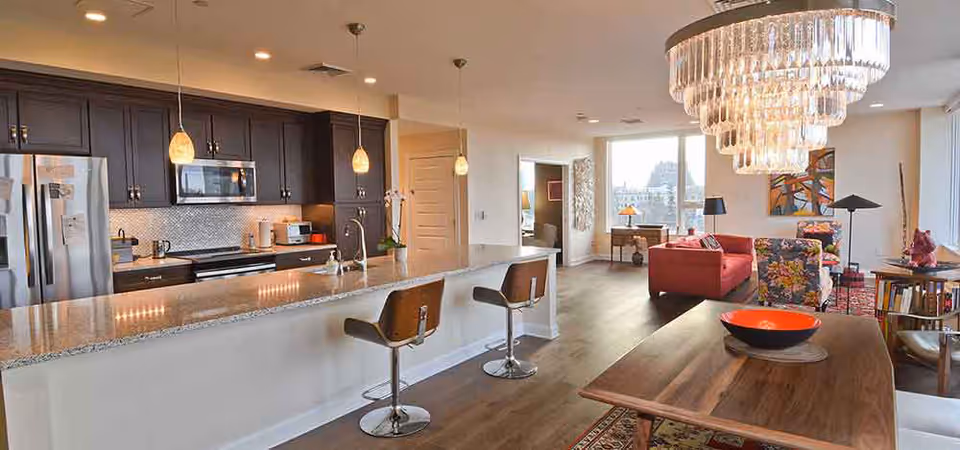 A modern open-concept living space featuring a kitchen with dark cabinetry, stainless steel appliances, and a granite countertop with two bar stools. The area extends into a living room with a red sofa, a floral armchair, a wooden dining table with a decorative bowl, and a large chandelier. Large windows provide natural light and a view outside.