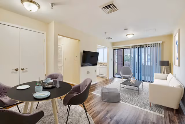 A modern living and dining area in a senior living facility with a round dining table set with plates and cups, four purple chairs, a wall-mounted flat-screen TV, a white sofa, a gray armchair, a coffee table, a floor lamp, and sliding glass doors with vertical blinds leading to an outdoor space.