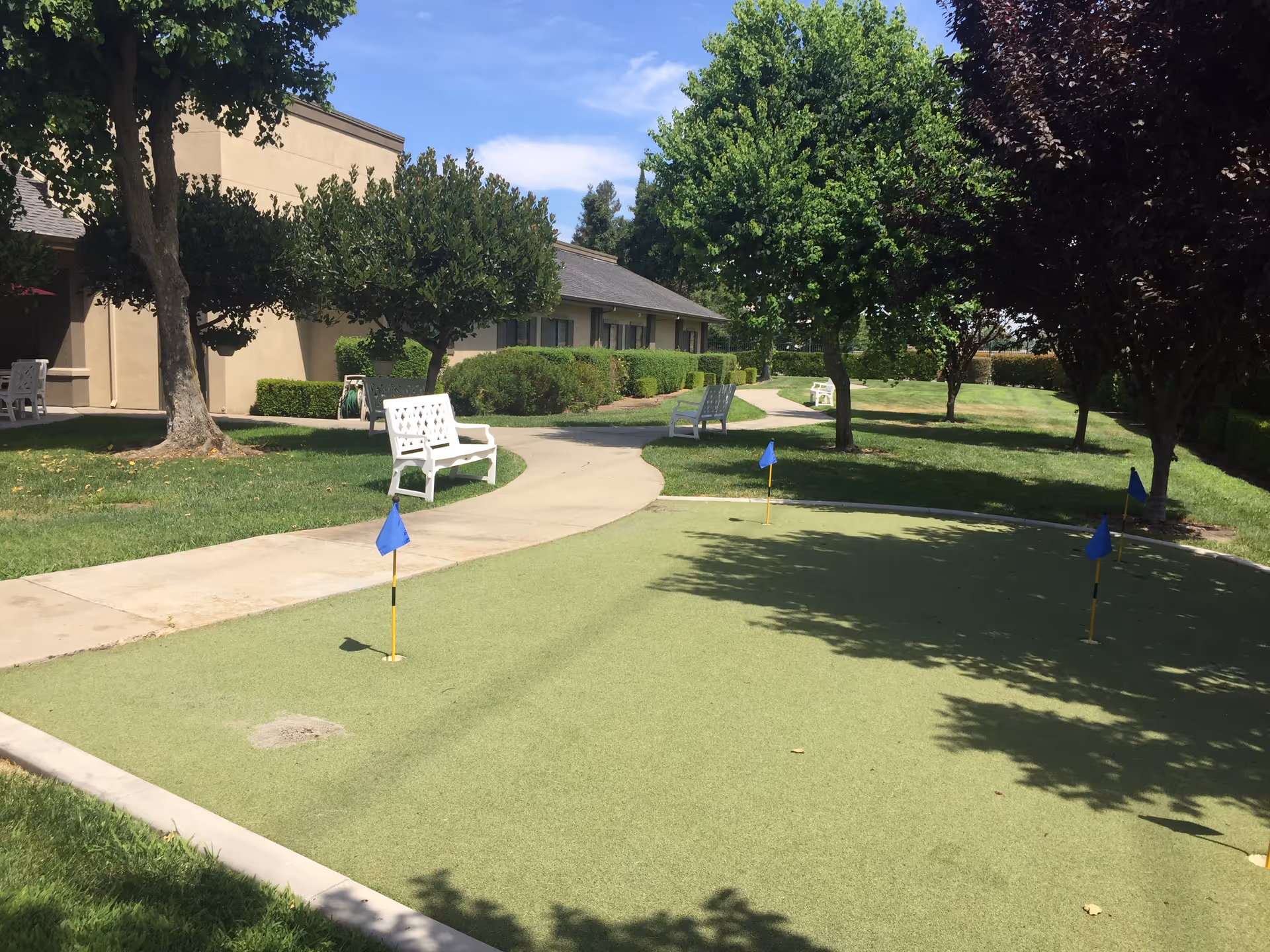 Outdoor putting green with four small blue flags, surrounded by a paved walkway, white benches, trees, and bushes near a beige building under a blue sky.