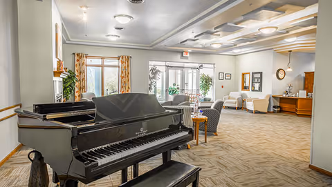 Spacious senior living common room with a black grand piano, multiple seating areas, and glass entrance doors in the background.
