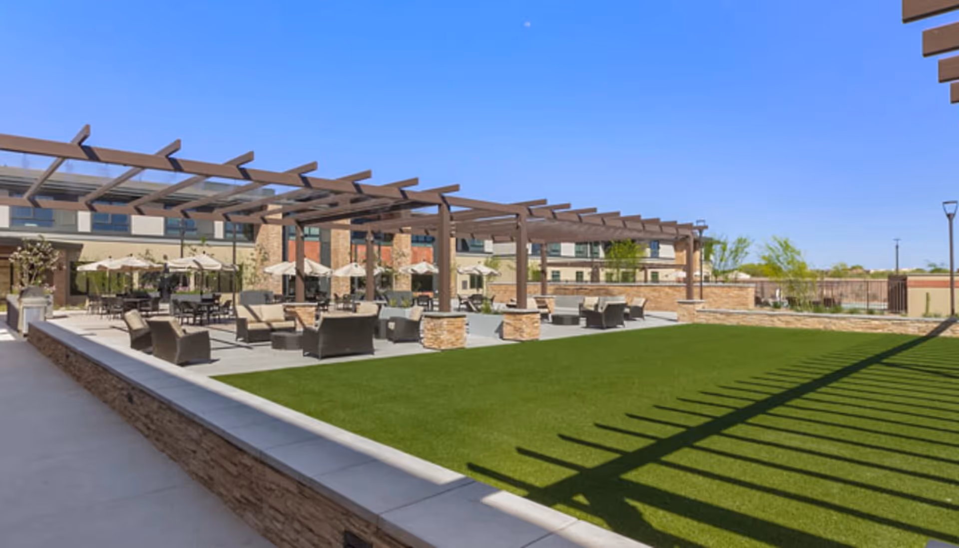 Outdoor patio area with seating arrangements including chairs and tables under large umbrellas and a wooden pergola structure, adjacent to a well-maintained green lawn, with a multi-story building in the background under a clear blue sky.