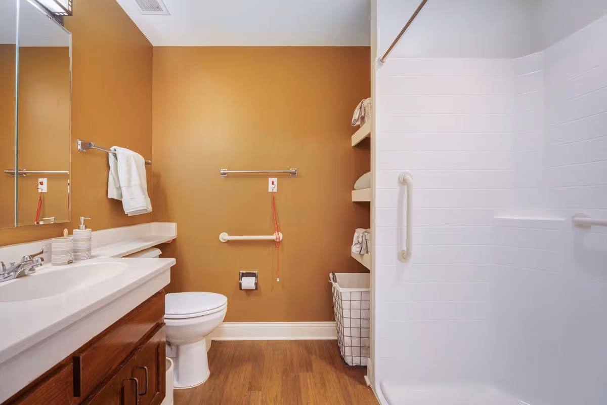 A clean and accessible bathroom with a white toilet, a wooden vanity with a white countertop and sink, a large mirror, and a walk-in shower with white tiled walls and grab bars. The walls are painted a warm tan color, and there are shelves with towels and a laundry basket next to the shower.