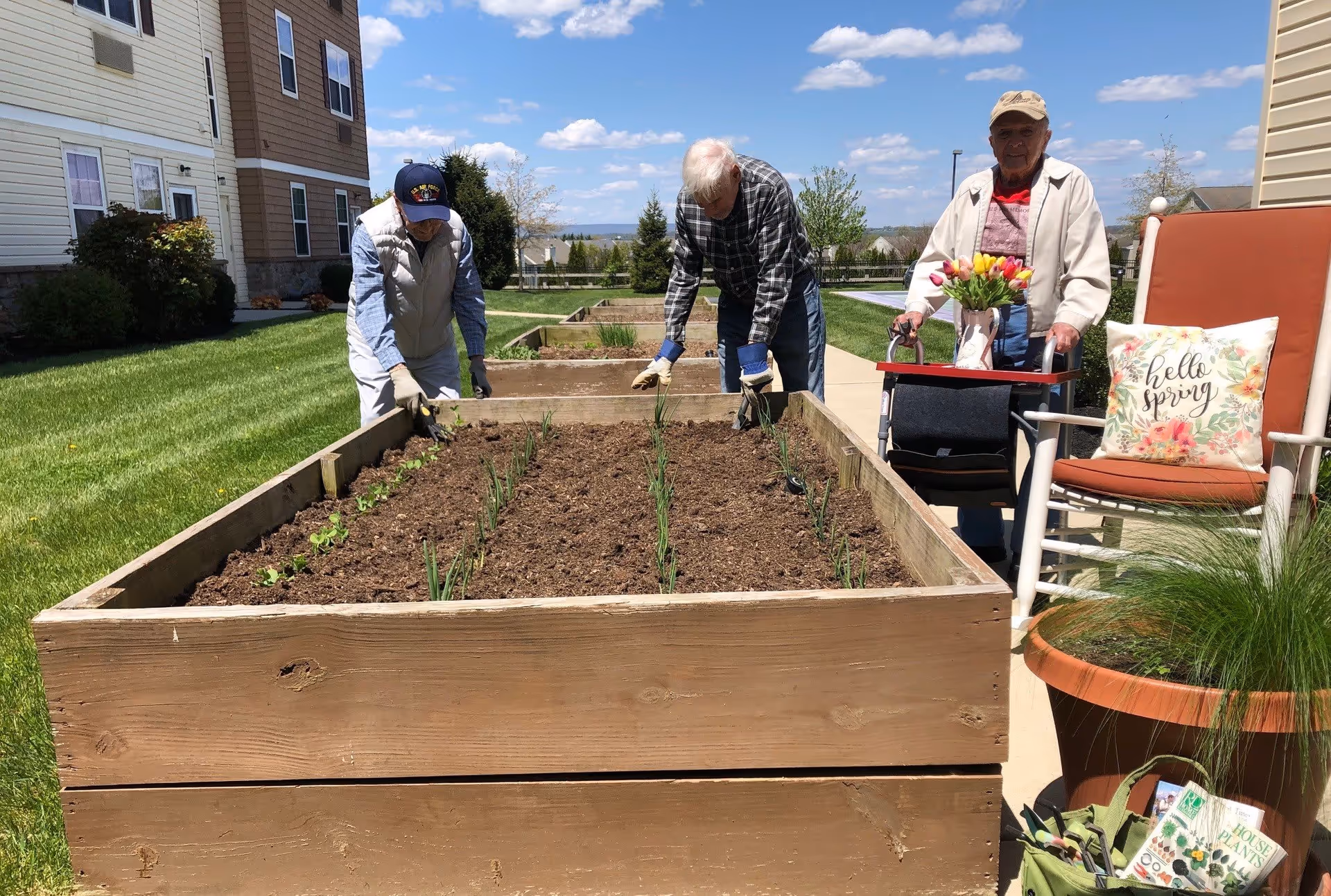 Three elderly individuals gardening in a raised wooden garden bed outside a residential building. Two men are tending to the plants in the garden bed, while a woman stands nearby with a walker that has a tray holding a vase of tulips. There is a cushioned chair with a floral pillow that says 'hello spring' and a large potted plant next to it. The scene is sunny with a blue sky and some clouds.