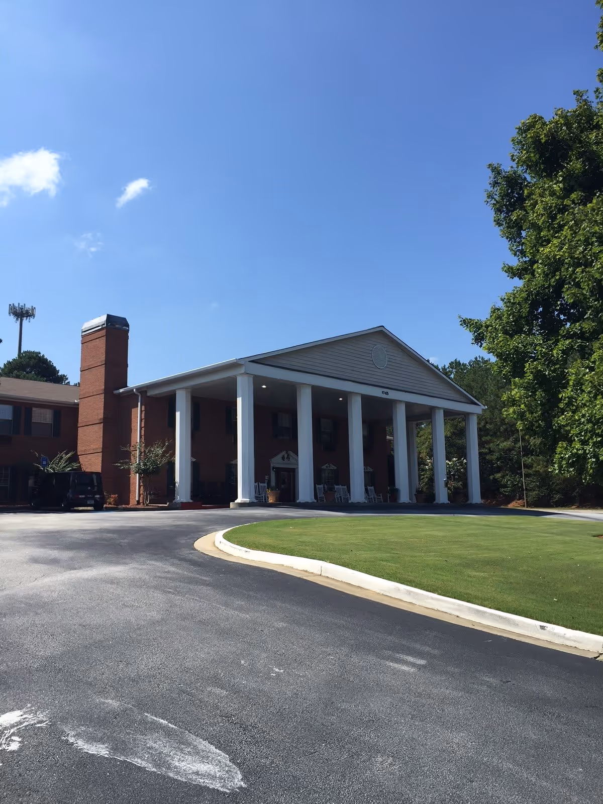 Front entrance of a brick senior living building with large white columns, a covered porte-cochere, driveway and lawn under a clear blue sky.