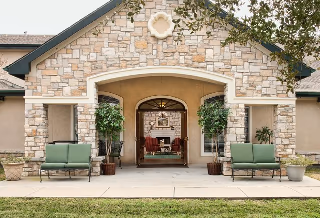 Stone-faced building entrance with an arched doorway flanked by green cushioned benches and potted plants, showing seating inside.