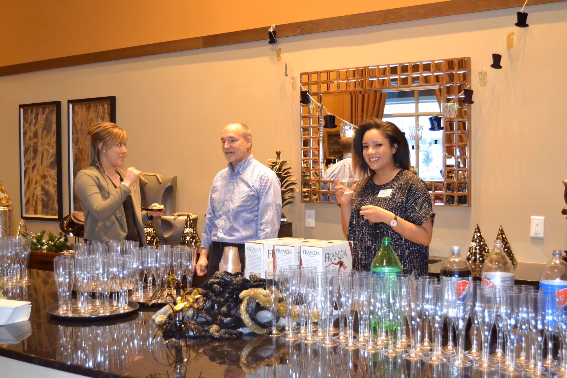 Three people standing behind a counter filled with empty wine glasses and bottles, including boxed wine and soda bottles. The woman on the right is holding a glass and smiling, while the other two people are engaged in conversation. The background features a large decorative mirror and wall art.