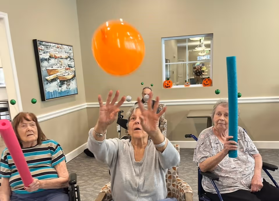 Three elderly women seated in a room, two holding foam pool noodles and one reaching up to catch an orange ball in mid-air. The room has beige walls, a painting of boats, and a window with Halloween decorations including two pumpkin faces and an owl.