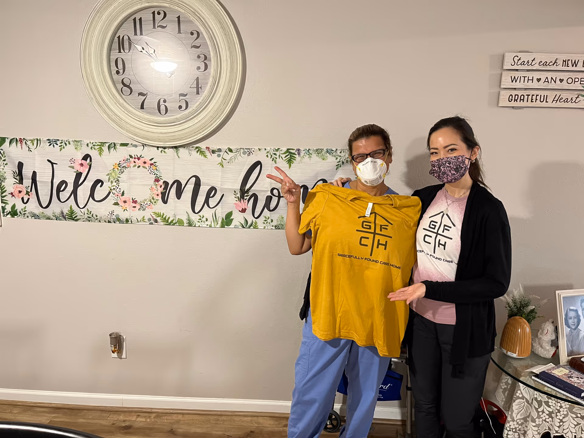 Two women wearing face masks stand indoors in front of a wall with a large round clock and a floral 'Welcome home' banner. One woman holds up a yellow T-shirt with a logo and text that reads 'Gracefully Found Care Home'. The other woman gestures towards the shirt. There is a small table with framed photos and decorative items to the right.