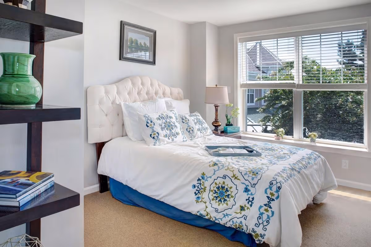 Bright bedroom featuring a tufted white headboard, patterned bedding, a bedside table with lamp, and a large window.