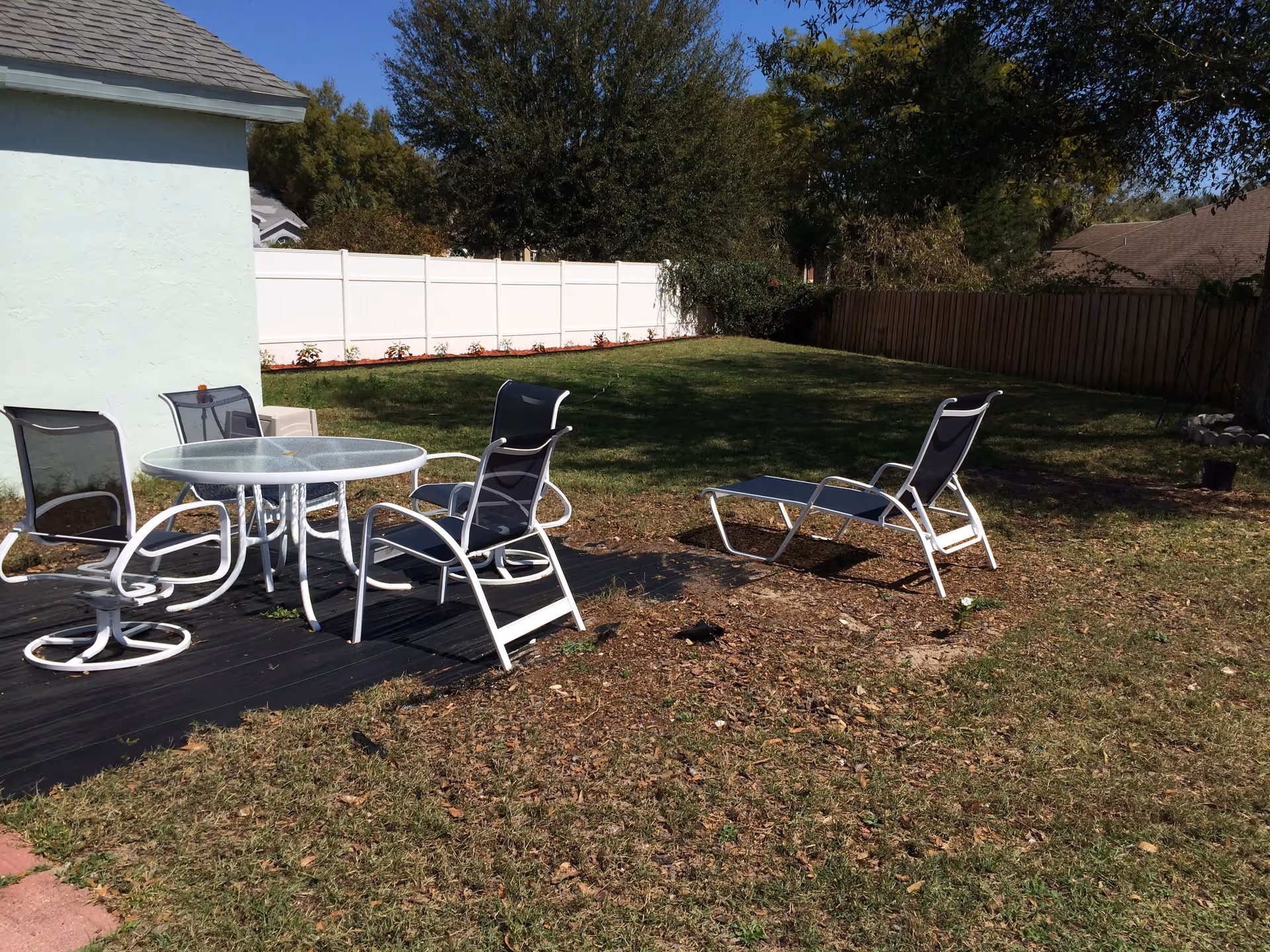 Outdoor backyard area with a glass-top round table and four white-framed chairs with black mesh seats. The area is adjacent to a light green building with a shingled roof. The yard is grassy with a white fence on one side and a wooden fence on the other, surrounded by trees and shrubs under a clear blue sky.